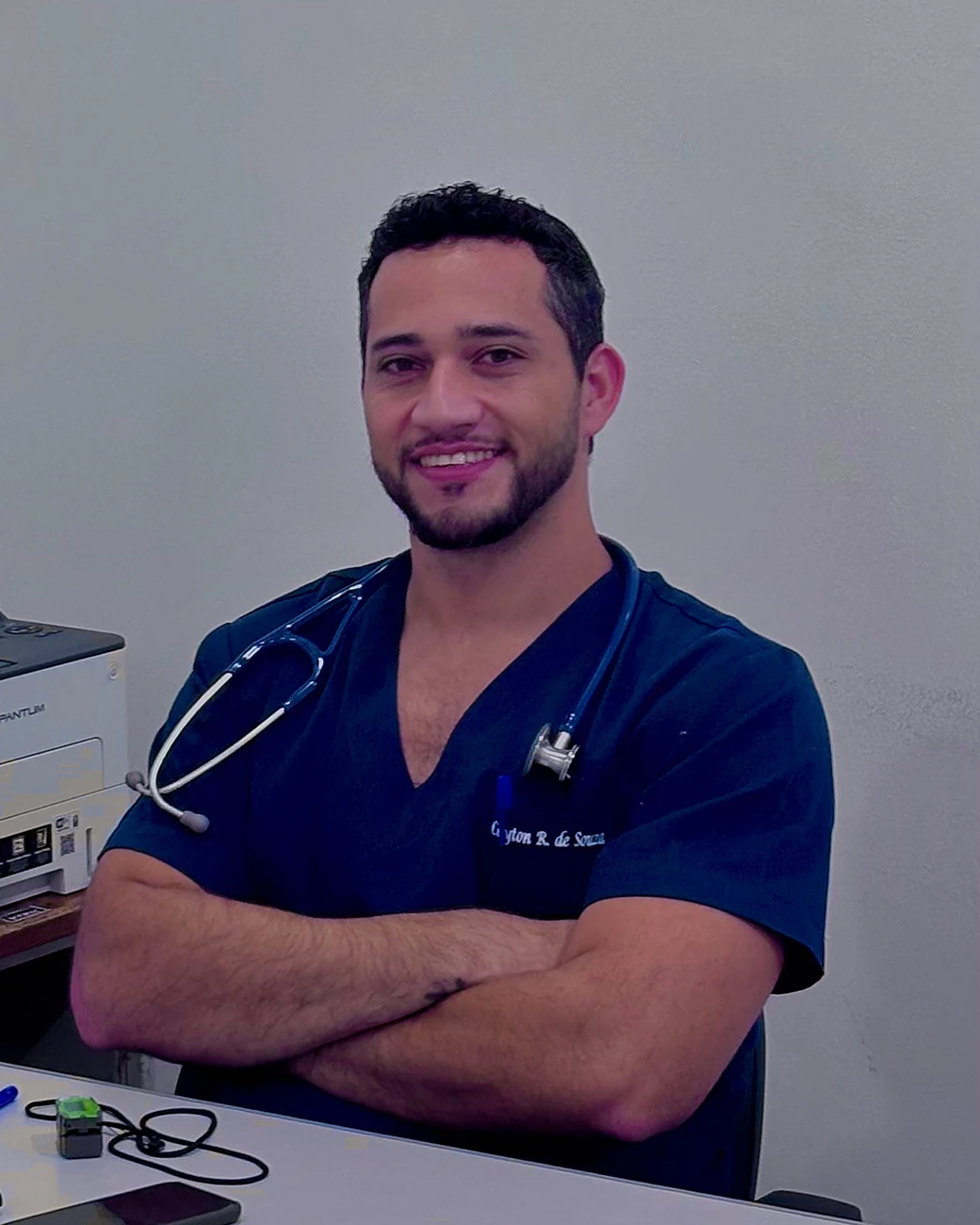 Dr. Cleyton sentado à mesa do consultório, de braços cruzados e sorrindo